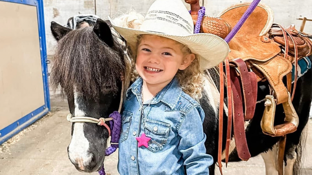 A young girl in a tan straw cowgirl hat stands smiling with a small pony. On the pony's back is a tan leather saddle with stirrups.