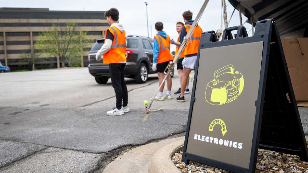 A tall sign reads "Recycling Electronics" and features a CD Player. Behind the sign is a parking lot with four teenage boy volunteers standing in bright orange construction vests.
