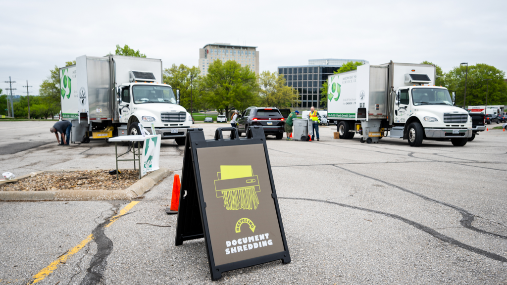 A sign that reads "Recycling Document Shredding stands in a parking lot at Recycling Extravaganza. Behind the sign sits large white trucks and volunteers helping to shred papers.